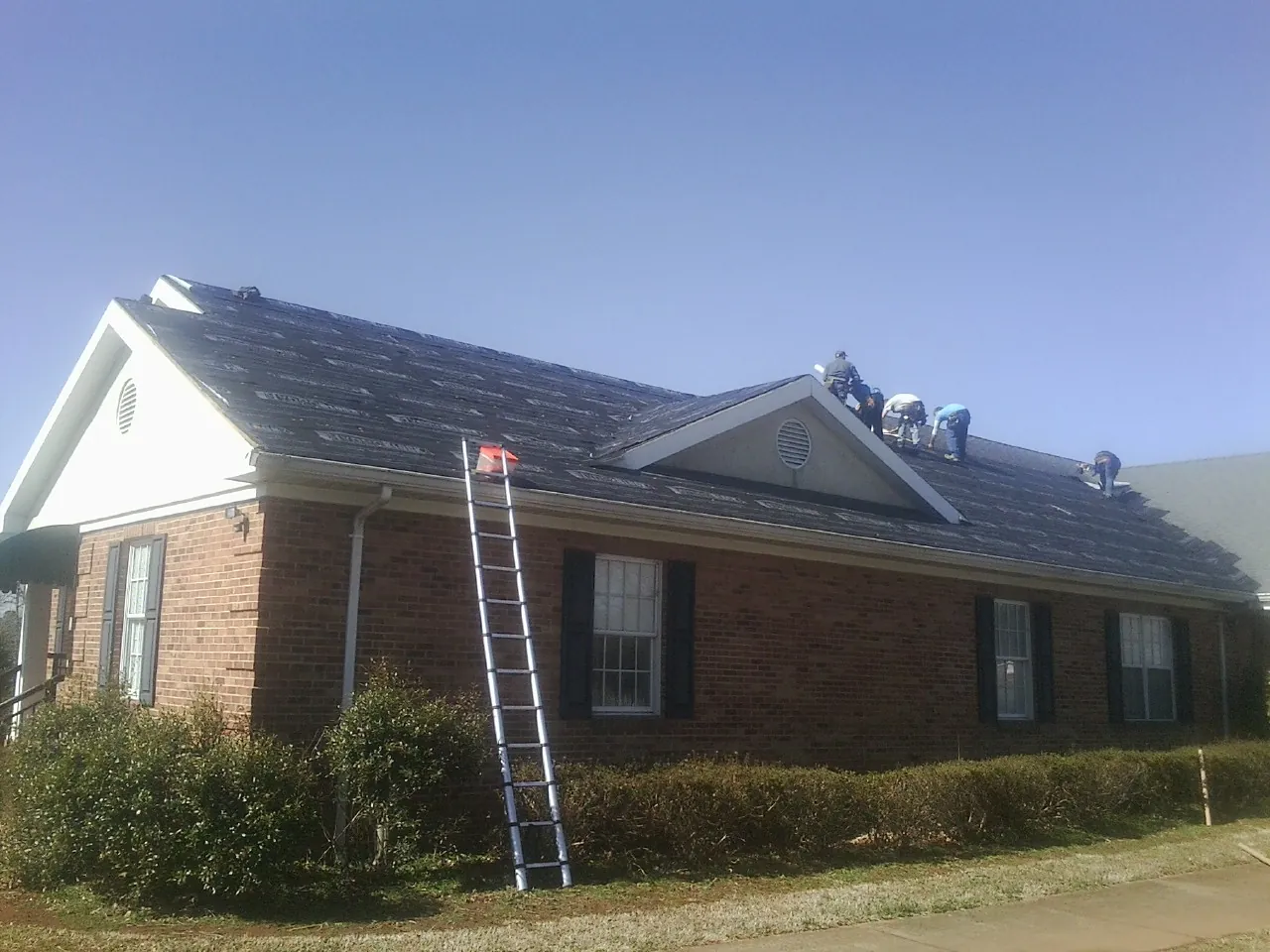 Crew tearing shingles off a brick ranch roof with a ladder set — Emergency Roof Repair work by our crew in Fort Gaines