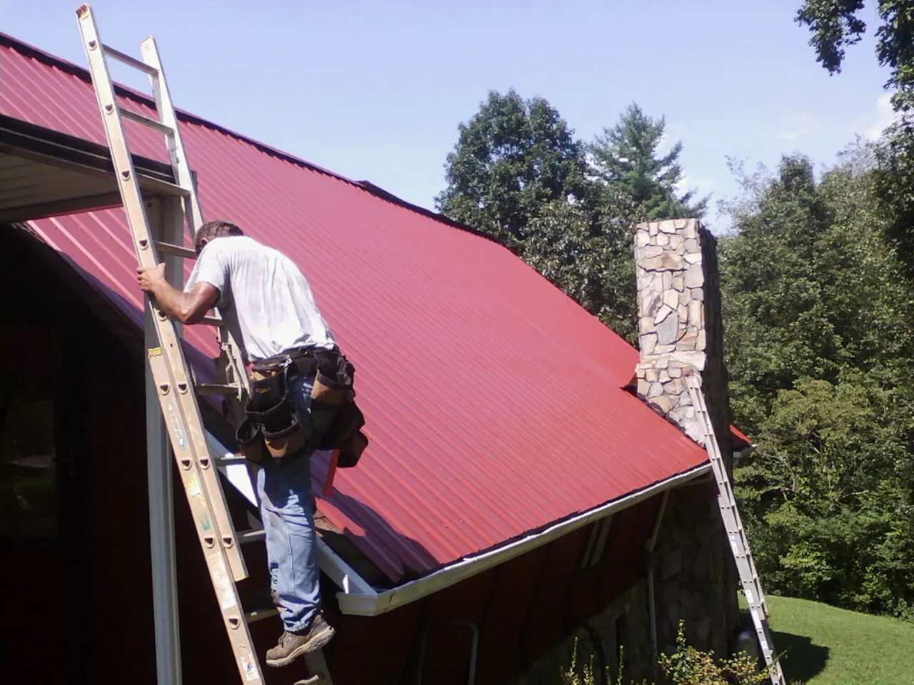 Roofer on a ladder beside a red metal roof and stone chimney — Soffit & Fascia Repair by Roofing Brothers in Fort Gaines